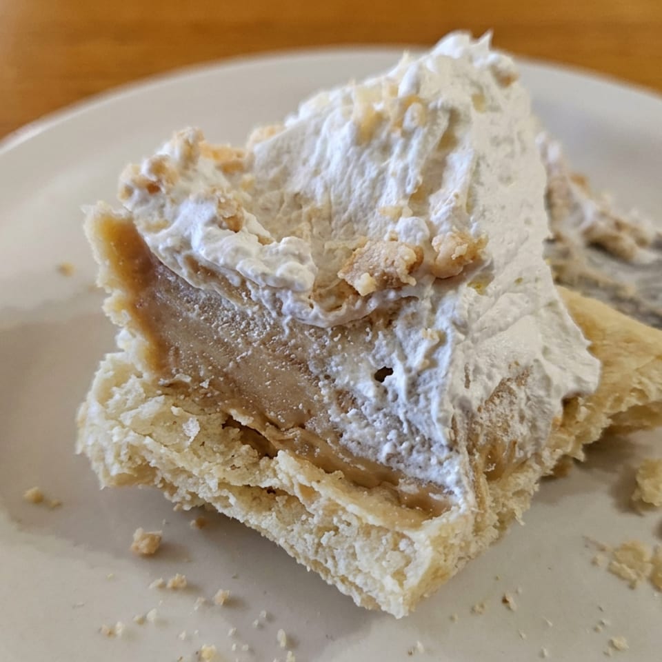 Slice of crumbly pie topped with whipped cream and caramel filling on a white plate atop a wooden table.