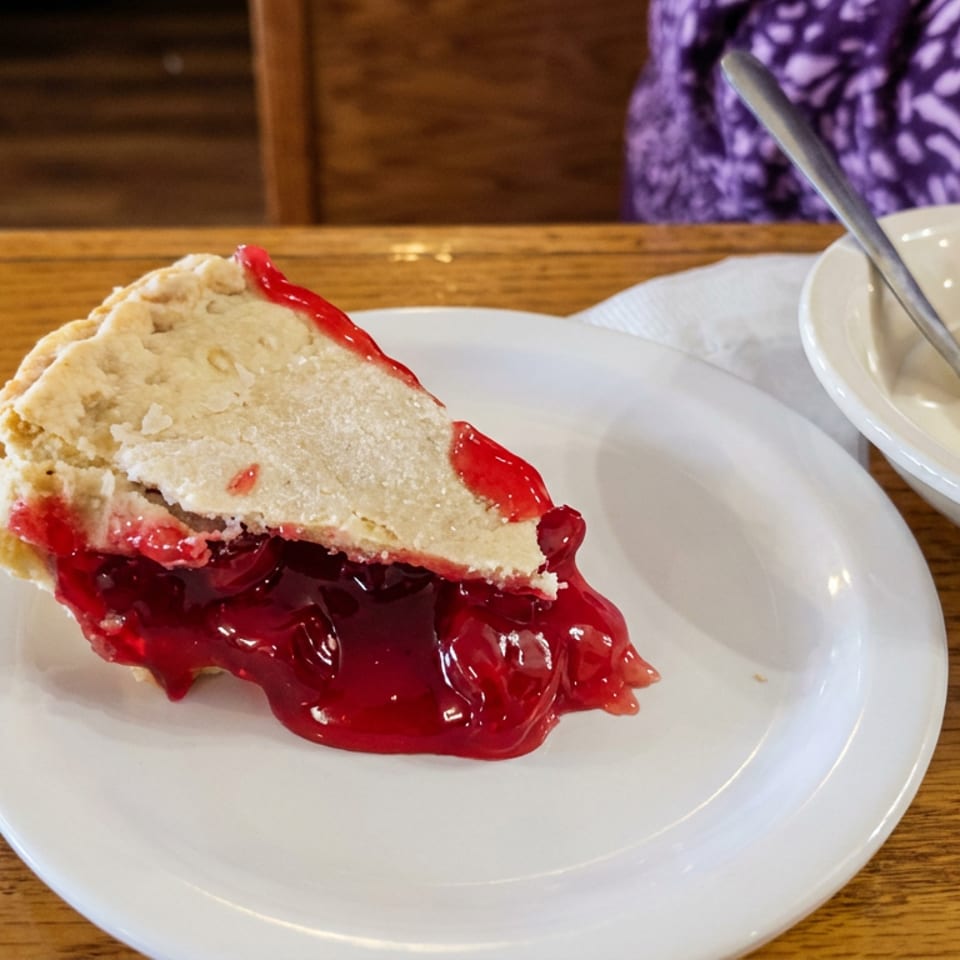 Slice of cherry pie with flaky crust and whipped cream on a white plate at a wooden restaurant table.